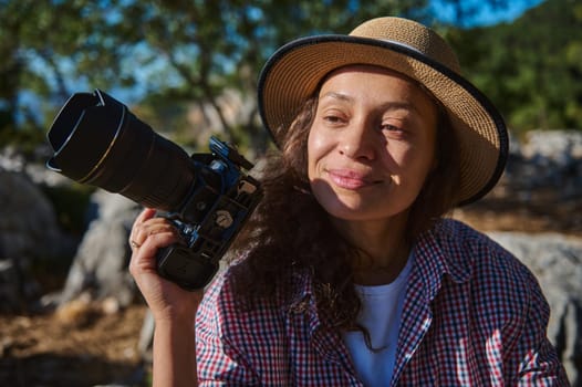 A joyful woman in a hat holds a camera, basking in sunlight. She appears content, surrounded by nature's beauty, capturing serene moments. Ideal for concepts of travel, photography, and happiness.