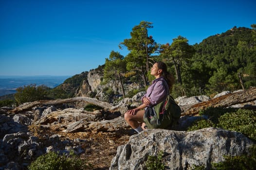 A woman sits peacefully on rocky terrain, taking in the beautiful mountain scenery around her. The sun shines brightly, creating a perfect atmosphere for reflection and tranquility during a hike.