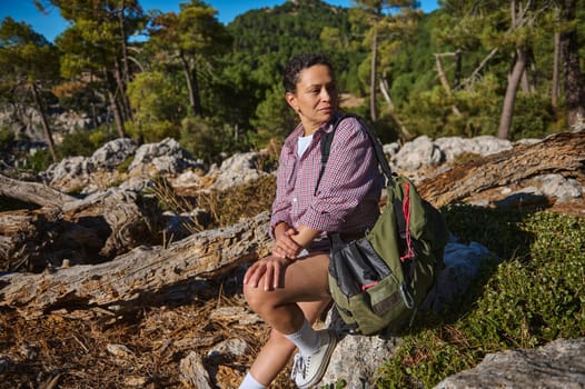 A woman sits on fallen logs in a forest, enjoying a peaceful hiking break. She wears casual outdoor attire, surrounded by lush greenery and rocky terrain, embodying tranquility and adventure.