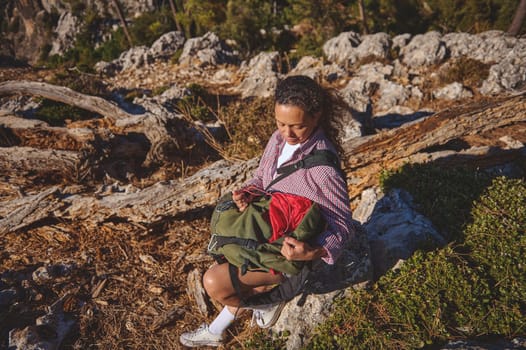A woman enjoys a peaceful moment in nature, sitting on a rock while checking her backpack during a hike in a rugged, rocky landscape surrounded by trees.