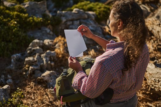 A woman in a plaid shirt examines a piece of paper while hiking through a rugged, rocky terrain. The scene conveys a sense of exploration and discovery amidst an adventurous outdoor environment.
