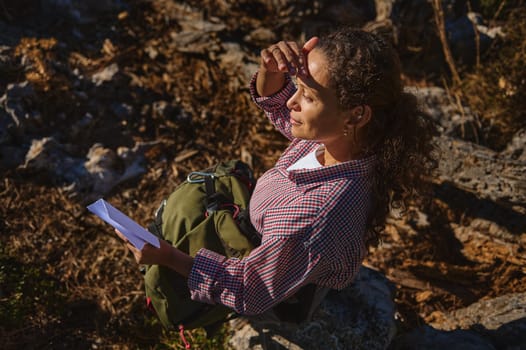 A woman with curly hair sits on rocks in the outdoors, holding a map. Dressed in a checkered shirt, she shades her eyes from the sun, suggesting a hiking or exploring adventure.