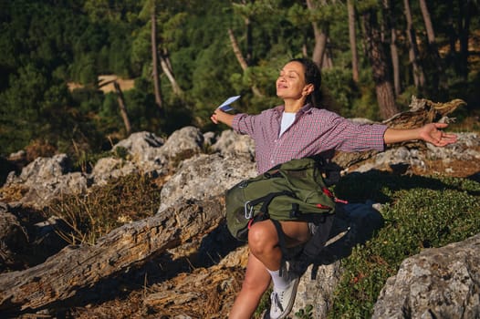 A woman sitting on rocks with arms outstretched, embracing the sunshine in a serene forest environment, symbolizing freedom and relaxation.