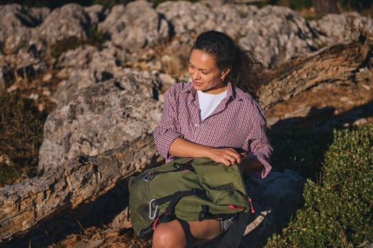 A woman relaxes on a rocky outdoor landscape, enjoying the sunlight and nature. She sits with a green backpack, wearing a casual checkered shirt. Sunlight enhances the peaceful mood.