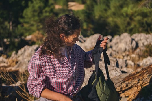 A woman wearing a plaid shirt stands with a backpack in a rocky outdoor area, surrounded by nature and sunlight.
