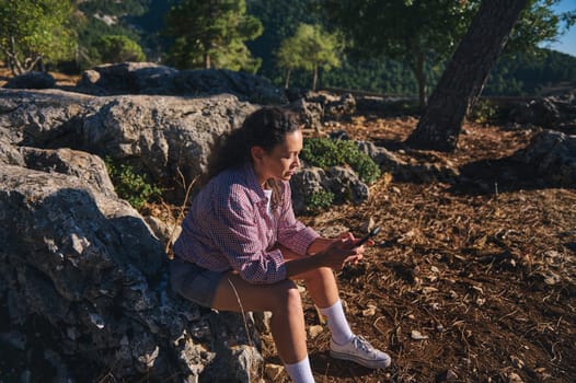 A woman sits on rocky terrain, engrossed in her smartphone, surrounded by nature. The scene depicts leisure and technology blending in a tranquil outdoor setting.