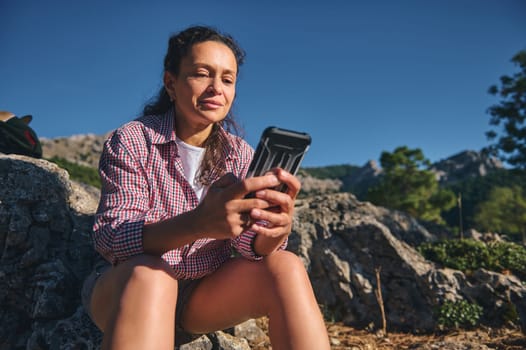 A woman in casual attire sits on rocky terrain, enjoying the sun and using her smartphone. The backdrop features scenic mountains and clear blue skies, evoking calm and connection to nature.