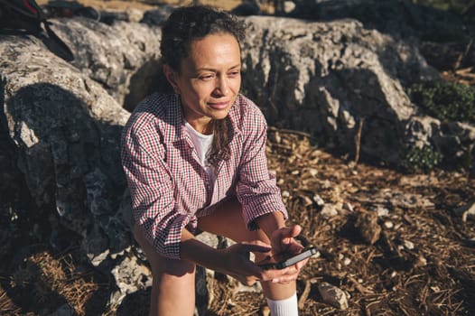 A woman in a checkered shirt sits on rocks outdoors, holding a phone. She appears relaxed and thoughtful, basking in warm sunlight.