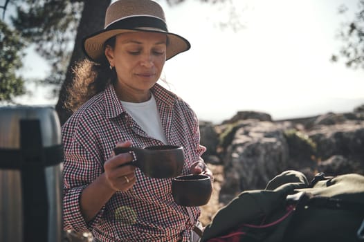 A woman in a hat enjoys a coffee break in nature, holding a cup while outdoors. Surrounded by trees and rocks, she exudes relaxation and serenity. Ideal for topics on travel and leisure.