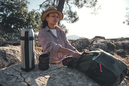 A woman relaxes outdoors with a thermos and backpack on a rock. The tranquil scene suggests a peaceful picnic amidst nature, perfect for enjoying solitude and tranquility.