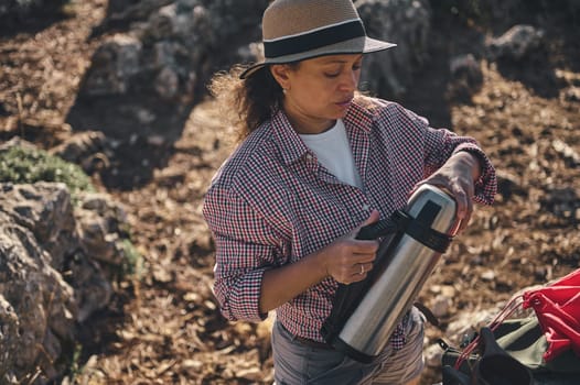 A woman in a checked shirt and hat pours a hot drink from a thermos during a hiking adventure. She seems relaxed and content, embracing the tranquility of nature.