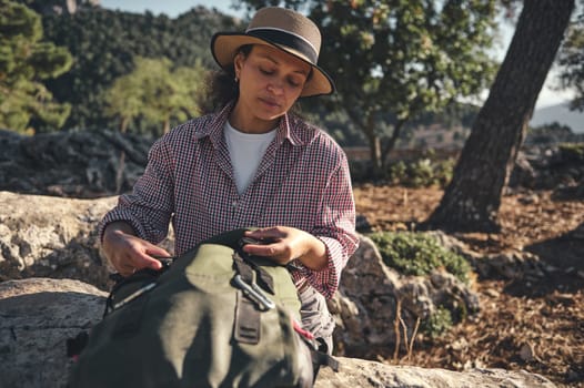 A woman in a sun hat and checked shirt sits outdoors with a backpack. She enjoys the tranquil natural surroundings, soaking in the serene atmosphere of the forest setting.
