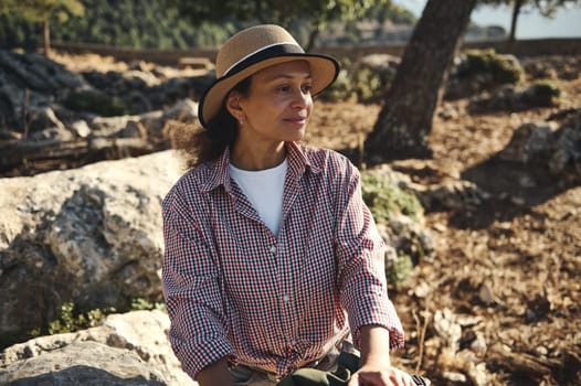 A woman sits on rocky terrain, wearing a checkered shirt and straw hat, enjoying a serene outdoor atmosphere. Natural beauty and relaxation are key themes, embraced by sunlight and nature.