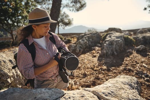 A woman wearing a hat explores a rocky landscape, carrying a backpack and camera. She enjoys nature and adventure under the clear sky, capturing moments while hiking in the outdoors.