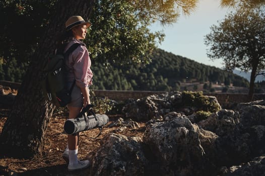 A woman rests under a tree with a backpack and yoga mat in a serene outdoor setting. She is surrounded by rocky terrain and lush forests under a sunny sky, conveying adventure and tranquility.