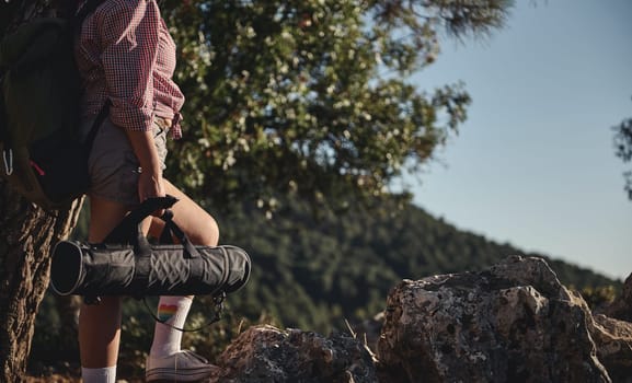 A hiker pauses on a rocky trail, carrying a yoga mat, surrounded by lush green forest. The scene captures the essence of outdoor adventure and mindfulness in nature.