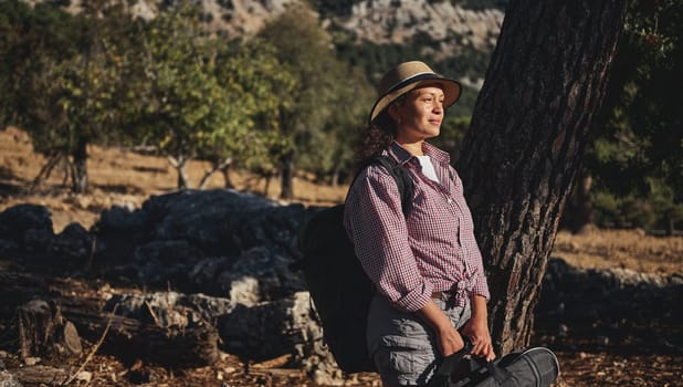 A woman standing under a tree with closed eyes, enjoying nature's tranquility. She is wearing a hat and backpack, surrounded by a sunny forest with a peaceful atmosphere.