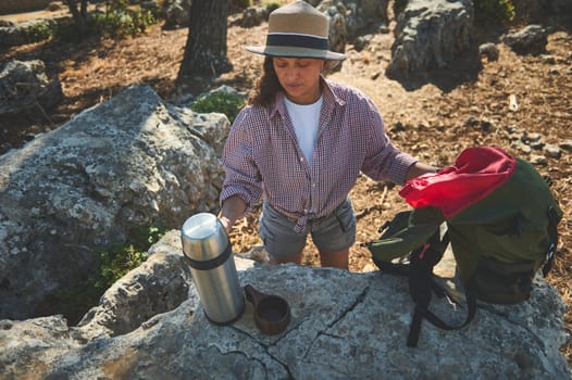 A woman in a hat takes a break during a hike, sipping a drink from a thermos on a rocky trail.