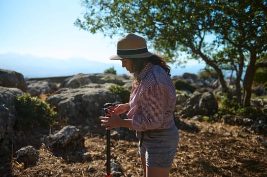 A woman wearing a hat adjusts a tripod in a sunlit rocky outdoor setting surrounded by greenery.