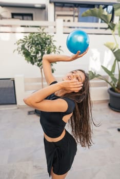 A joyful woman playfully poses with a bright blue ball in a sunlit, vibrant outdoor environment full of life
