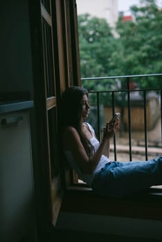 A woman sits by the window with her phone, surrounded by lush greenery in a peaceful, serene moment