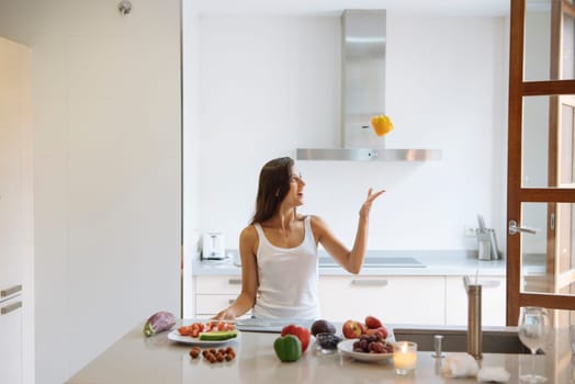 A woman is playfully juggling various citrus fruits in a modern kitchen filled with fresh, vibrant produce