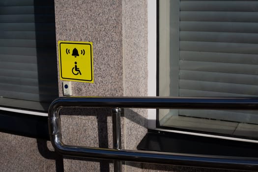 Hand rails to assist person in wheelchair next to handicap sign on wall and call button to assist person with disability, modern entrance to building with convenient ramp for people with disabilities