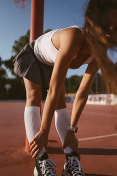 In a bright and lively outdoor basketball environment, a young athlete is tying her shoes with focus and enthusiasm
