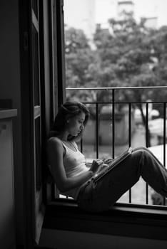 A woman sits comfortably in a cozy window nook, deeply reflecting while writing her thoughts in a notebook