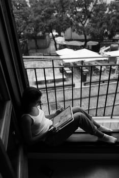 A tranquil black and white photograph showcasing a young woman deeply engrossed in a captivating book by the window