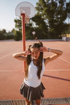 A cheerful teenage girl poses playfully on a sunny basketball court, showing her vibrant energy and style