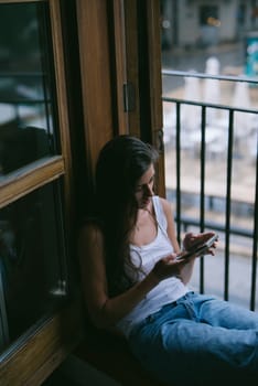 A young woman relaxes by a window, absorbed in her smartphone as rain creates a soothing atmosphere