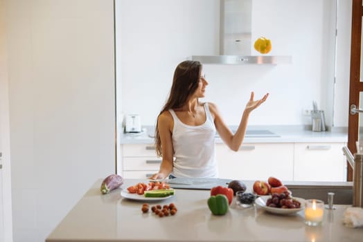 A woman is playfully juggling vibrant fruits in a bright, modern kitchen that is filled with fresh produce