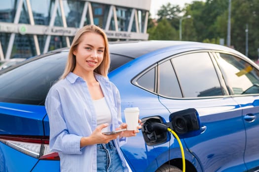 Blonde woman with long hair enjoys coffee while charging her electric car at a charging station outdoors.