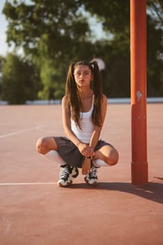 A young girl squats playfully on a basketball court, enjoying a carefree moment under the warm sunlight