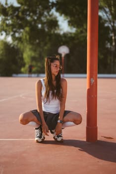 A confident young woman is squatting on a vibrant basketball court, showcasing her sporty style and urban vibes
