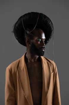Serious African American gay model wearing huge wig with chain over black background. Black man is wearing blazer in studio.