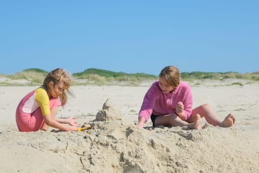 Two girls building a sandcastle on the beach under a clear blue sky. Two happy sisters having fun on the Atlantic ocean deserted beach.