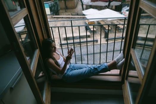 A young woman finds a serene moment of tranquility by the window, immersed in her phone while unwinding