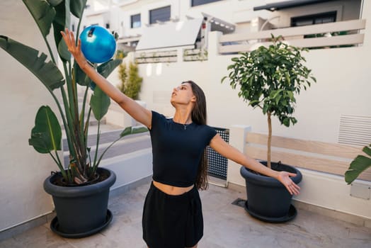 A woman is happily balancing a vibrant blue ball while striking a pose outdoors in a stylish and chic environment