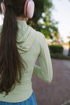 A young woman enjoys jogging outdoors, wearing a cozy hoodie and headphones, surrounded by greenery.