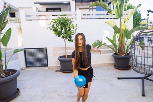 A young woman gracefully throws a vibrant blue ball in a stylish outdoor area that is surrounded by lush plants