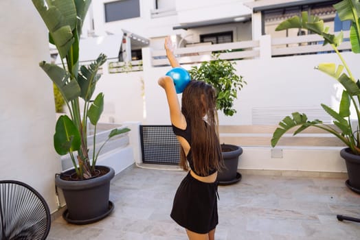 A young woman is exercising outdoors, holding a bright blue kettlebell while surrounded by lush greenery