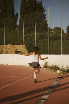 A spirited young girl joyfully twirls around on an outdoor sports court bathed in beautiful sunlight