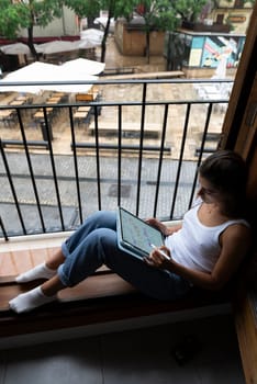 A woman is comfortably seated by the window, deeply engaged with her laptop in a calm and serene setting