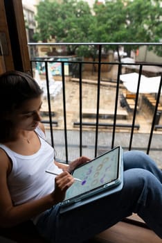 A young woman sits comfortably on her balcony, sketching creative ideas on her tablet while relaxing outside