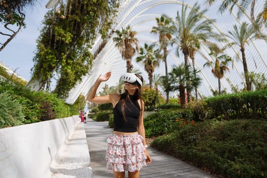 A woman waves cheerfully while standing on a pathway surrounded by palm trees and unique architecture.