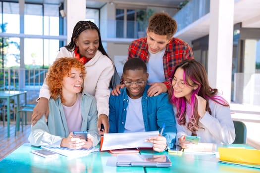 A group of five diverse students studying and collaborating at a library table working on a shared project, with books and devices on the table.