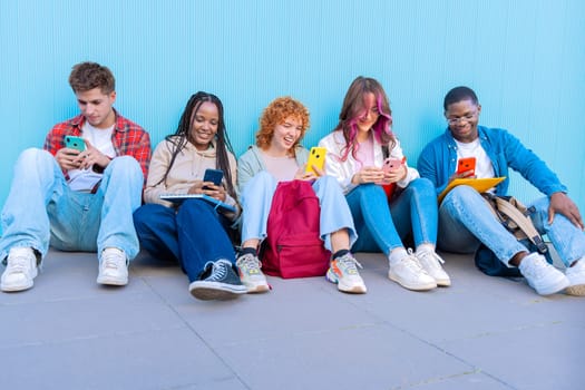 A diverse group of five smiling young students sit together on the floor, smiling and interacting on their smartphones against a blue background.