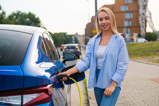 Portrait of blonde woman plugs in her electric car for charging in the street charging station.